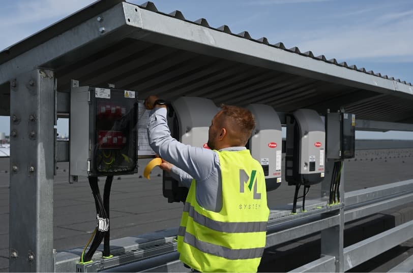 Technician in an ML System vest servicing rooftop solar inverters under a metal canopy.