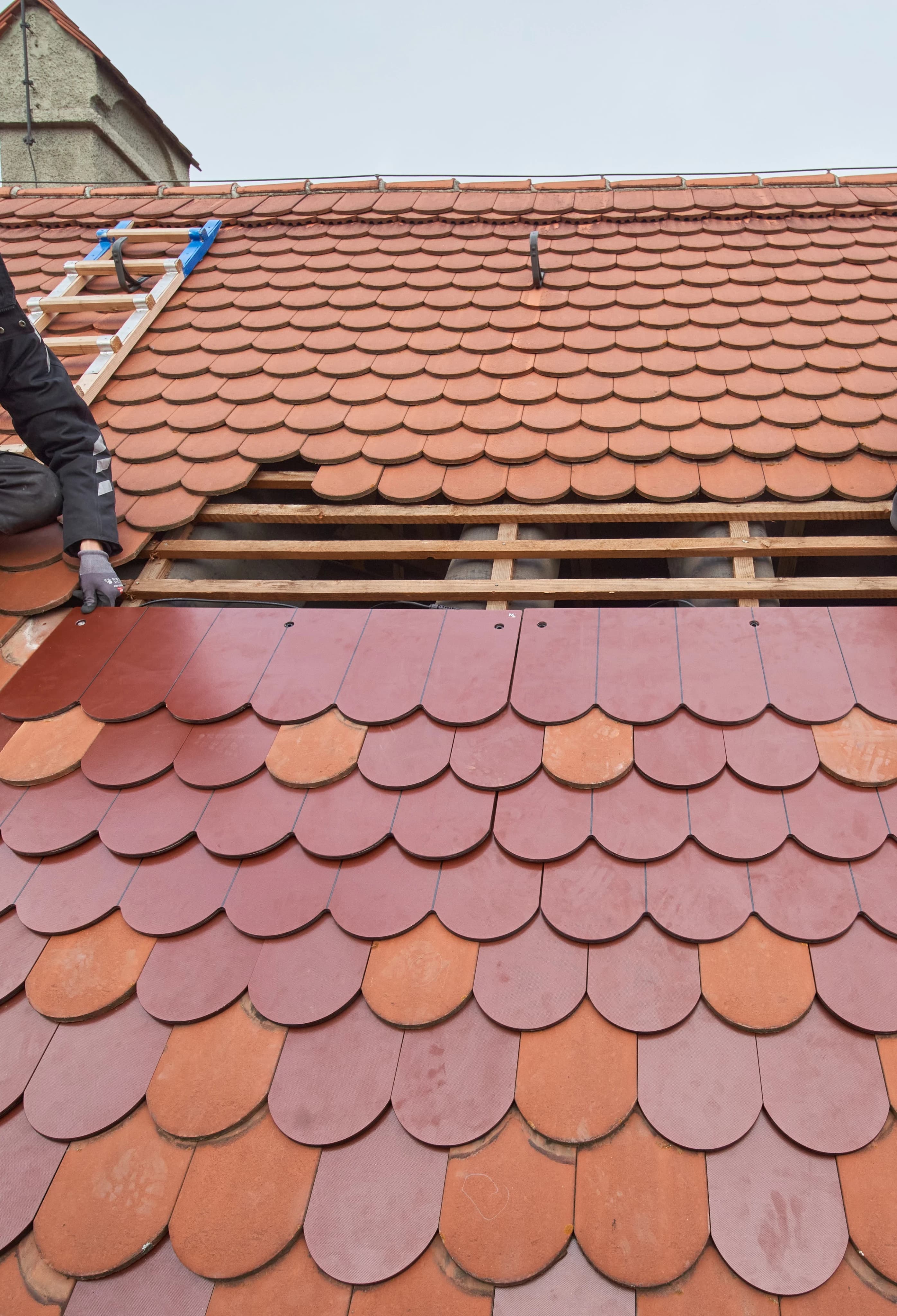A construction worker installs new reddish-brown fish-scale clay tiles on a sloped roof