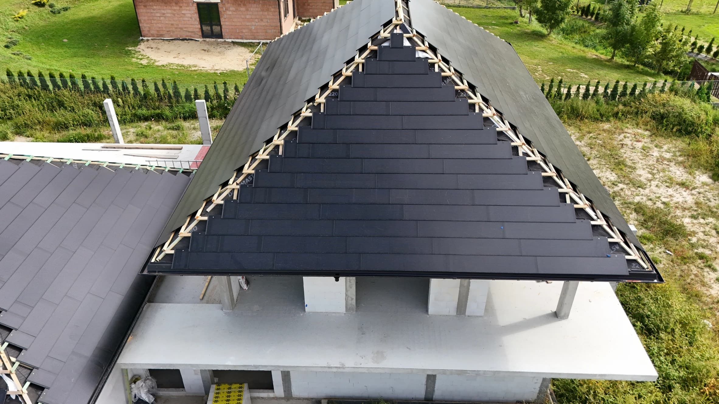 Aerial view of a modern house under construction with a dark, partially finished gable roof made of sleek solar tiles
