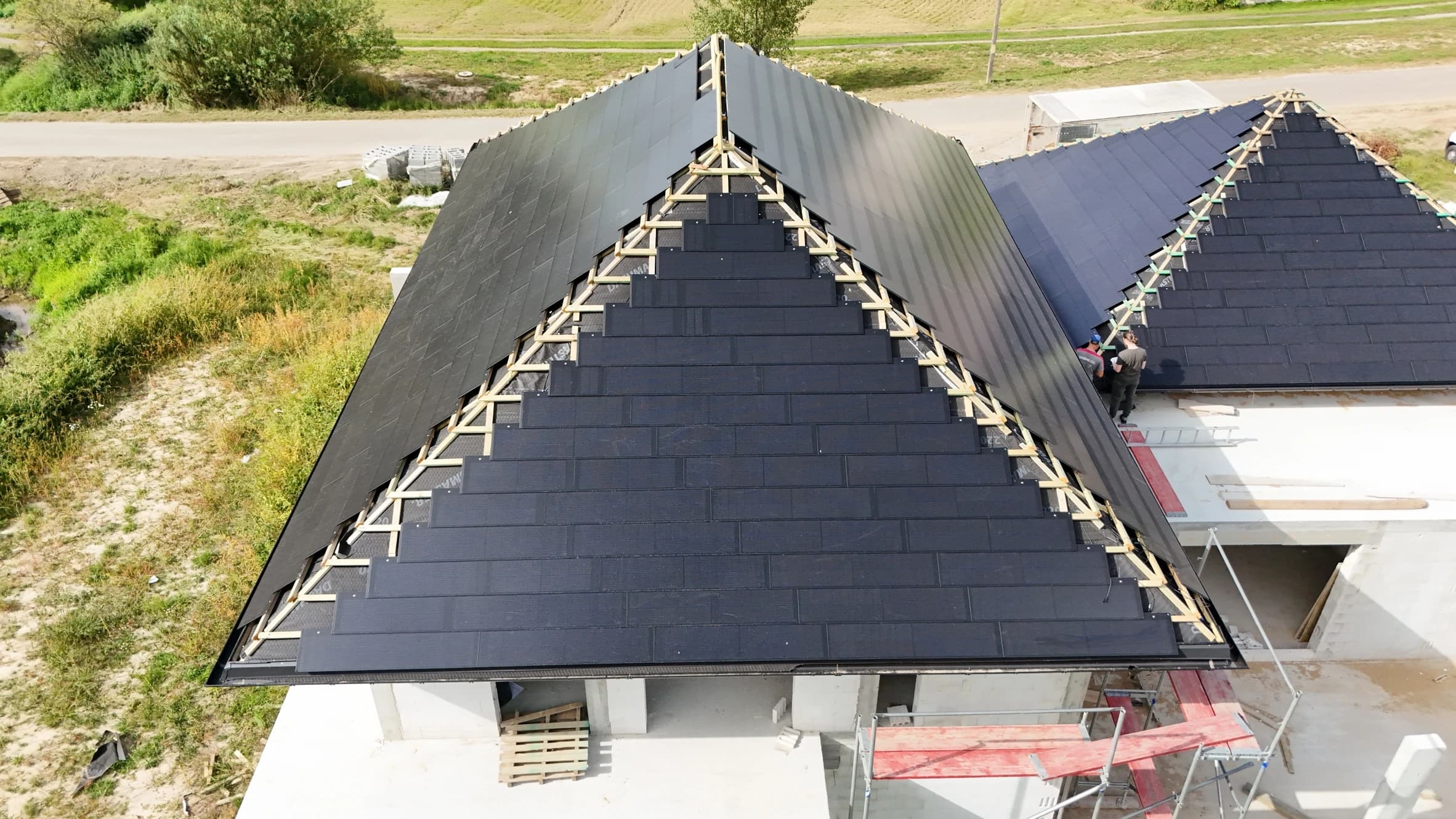 Aerial view of a house under construction with a dark, partially installed gable roof made of sleek solar tiles