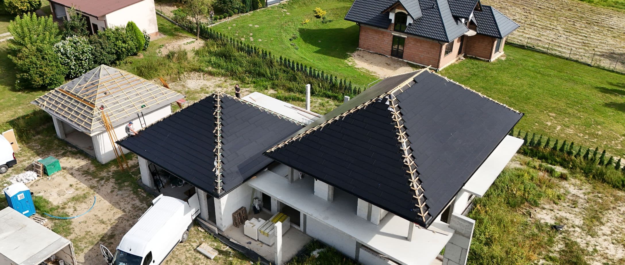 Aerial top view of a construction site featuring two adjoining buildings with dark, newly installed hip roofs made of photonroof tiles